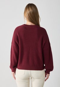 Burgundy knitted sweater with a textured finish, dropped shoulders, and long sleeves, paired with cream-colored pants.