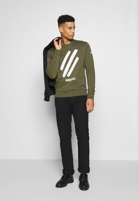 Felpa verde oliva con strisce grafiche bianche e testo, abbinata a pantaloni neri e scarpe nere. Il modello tiene in mano una giacca nera.