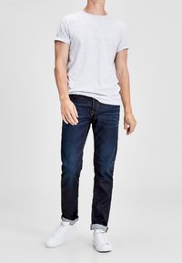 Camiseta de algodón gris, jeans oscuros y zapatillas blancas. La camiseta tiene un cuello redondo; los jeans son de corte recto con puños enrollados.