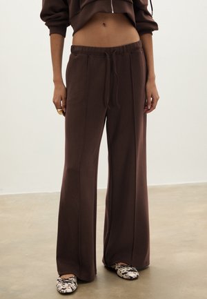 Pantalon marron à jambes larges avec coutures frontales, taille à cordon de serrage, porté avec un sweat à capuche marron court et des ballerines noires et blanches à motifs.