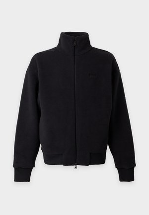 Un homme porte une veste noire à capuche et à fermeture éclair, un T-shirt gris, un pantalon noir et des chaussures noires, sur un fond blanc uni.