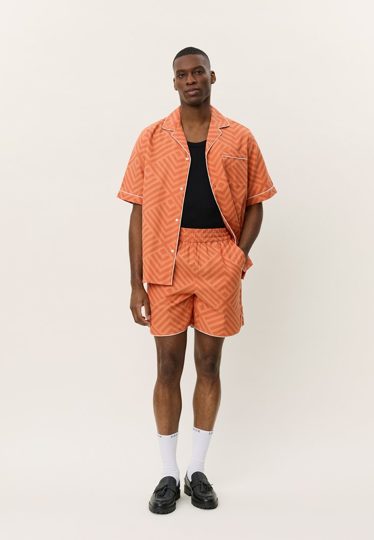 Camicia a maniche corte arancione con motivo e rifiniture bianche, abbinata a pantaloni corti coordinati. Indossata con una canotta nera, calzini bianchi e scarpe nere.