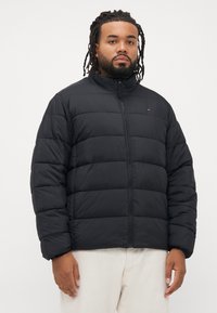 Mand med dreadlocks iført en sort pufferjakke og hvide bukser, stående mod en enkel lys baggrund.