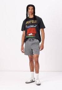 T-shirt graphique noir arborant un design de Snoop Dogg, associé à un short gris et des chaussettes de sport blanches. Les chaussures sont principalement blanches avec des accents noirs.