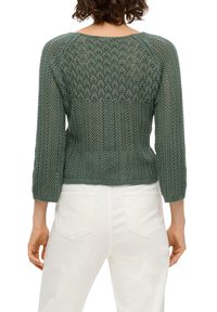 Grüner gestrickter Pullover mit Dreiviertelärmeln, der ein strukturiertes, offenes Strickmuster und einen gerippten Saum aufweist. Kombiniert mit weißen Jeans.