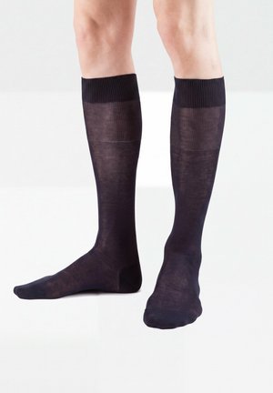 Beine, die durchsichtige schwarze kniehohe Socken tragen, stehen auf einer weißen Oberfläche, ein Fuß flach, der andere mit gespitzen Zehen.