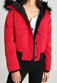 Červená vycpaná bunda s černým zipem, velkými kapsami a kapucí lemovanou kožešinou. Má elastické manžety a zkrácený střih.