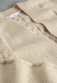 Béžový pletený sveter s textúrovaným dizajnom, vybavený rebrovaným golierom a manžetami, s hladkým a jemným povrchom. Vo vnútri viditeľná značka.