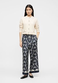 Γυναίκα όρθια με τα χέρια στις τσέπες, φοράει κρεμ ριγωτό ζακέτα με κουμπιά, φαρδιά μπλε σκούρα παντελόνια με λευκό ανθισμένο σχέδιο και μαύρες μπότες χωρίς τακούνι.