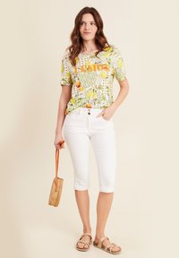 T-shirt met korte mouwen, geel en groen met een fruitpatroon, voorzien van oranje "LEMON" tekst, gecombineerd met witte capri-pantalon en bruine sandalen. Geweven handtas.