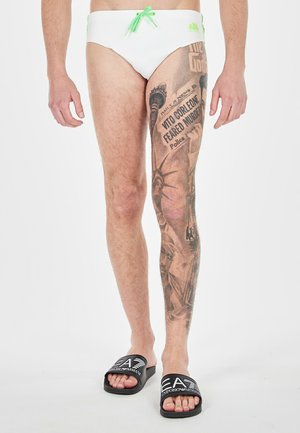 Pantaloni da bagno bianchi con cordoncino verde, caratterizzati da una gamba coperta di tatuaggi dettagliati, ciabatte nere con logo, texture della pelle liscia.