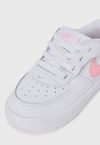 Sneaker Nike bianco con swoosh rosa, logo rosa sulla linguetta, lacci bianchi e punta perforata su sfondo bianco.
