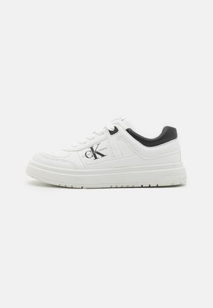 Fehér sneaker sima bőrből készült felsőrésszel, fekete sarokdíszítéssel, texturált gumi talppal, és az oldalán logóval, valamint kerek fűzőkkel rendelkezik.