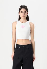 Bela rebrasta kratka majica brez rokavov s rožnato vezenim logotipom na sredini, kombinirana s črnimi hlačami z visokim pasom in srebrnimi kovinskimi detajli.