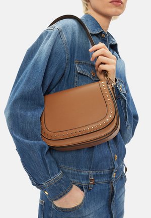 Persona che indossa una giacca di jeans blu e tiene una borsa a tracolla in pelle marrone con borchie e dettagli dorati.