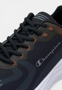 Musta urheilujalkine, jossa on sileä pinta, oransseja pistekoristeluita ja näyttävät nauhat, sekä harmaa Champion-logo sivulla.