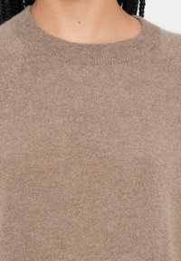 Μπεζ πλεκτό πουλόβερ με στρογγυλή λαιμόκοψη, ριμπ λαιμόκοψη και λεία υφή, παρουσιάζοντας μια κοντινή εικόνα του υφάσματος.