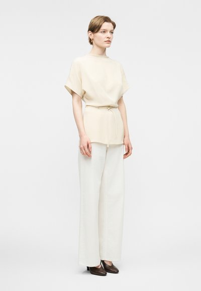 Femme debout portant un haut beige ample à manches courtes avec ceinture, un pantalon large blanc et des chaussures marron à petit talon, sur fond uni.