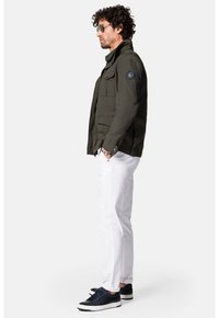 Oliwkowa kurtka z wieloma kieszeniami, zapinana na zamek, z naszywką logo na rękawie, w połączeniu z białymi spodniami i ciemnymi sneakersami.
