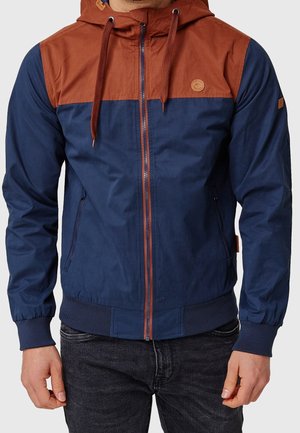 Mężczyzna ubrany w granatowo-rdzawobrązową kurtkę z kapturem, zapinaną na zamek z przodu i wyposażoną w sznurki, w zestawieniu z ciemnymi dżinsami.