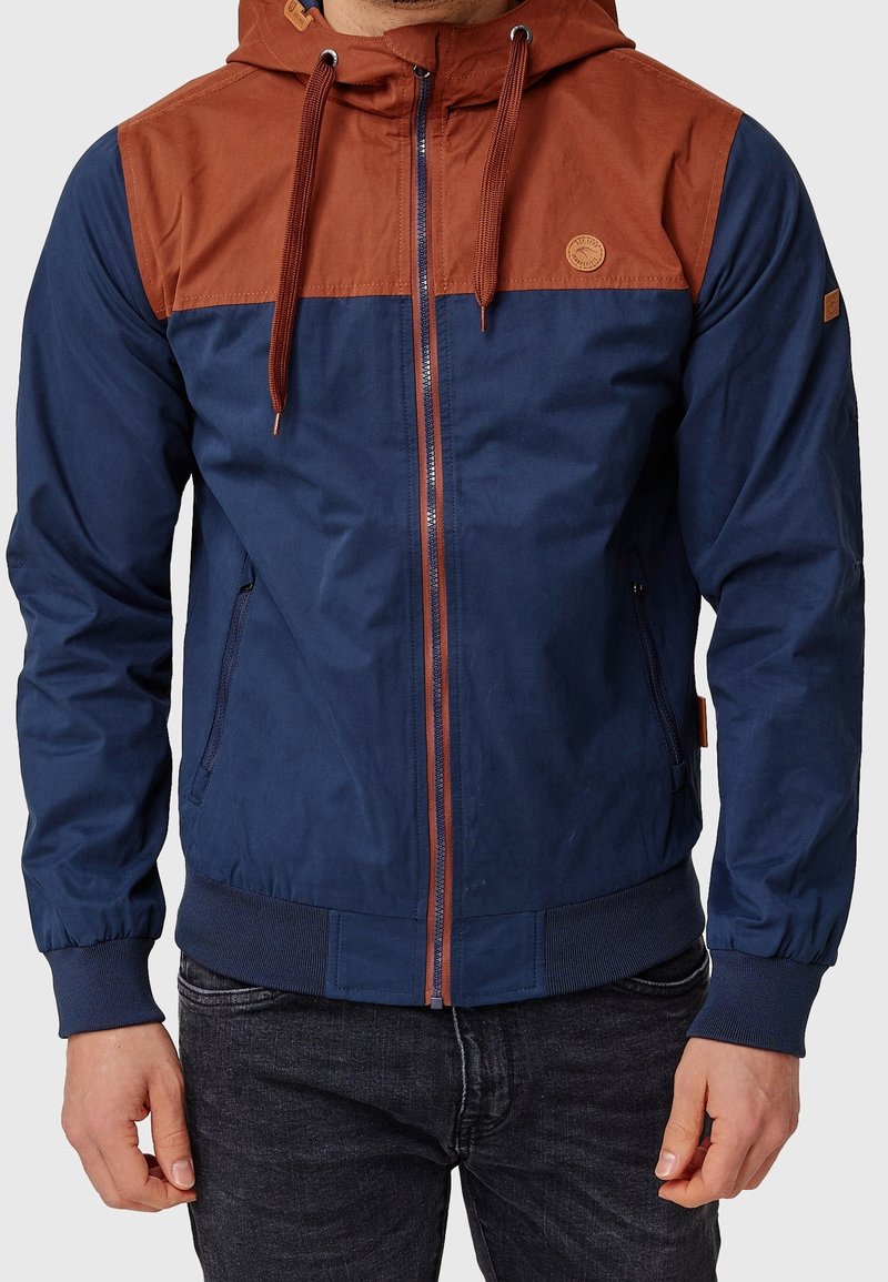 Homme portant une veste à capuche bleu marine et marron rouille avec une fermeture éclair à l'avant et des cordons de serrage, assortie à un jean foncé.
