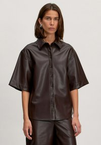 Braunes Kunstleder-Hemd mit kurzen Ärmeln, durchgehender Knopfleiste, spitzem Kragen und lockerer Passform. Glatte Textur und minimalistisches Design.