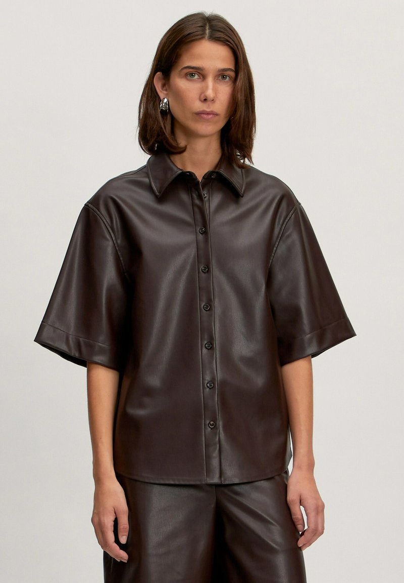 Braunes Kunstleder-Hemd mit kurzen Ärmeln, durchgehender Knopfleiste, spitzem Kragen und lockerer Passform. Glatte Textur und minimalistisches Design.