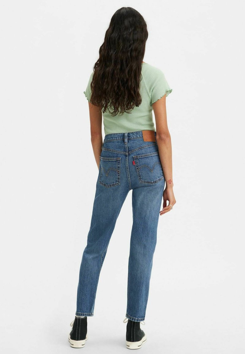 Haut tricot vert clair avec des bords en feston, associé à un jean en denim bleu coupe droite avec une patch en cuir et des poches arrière bien visibles.