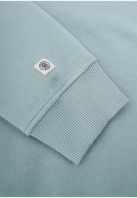 Vaaleansininen huppari, jossa on sileä pinta ja ribbauskankaiset hihansuut. Hupparissa on pieni brändilogo hihassa, joka osoittaa materiaalin ja suunnitteluasiat.