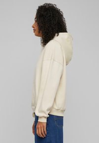 Lys beige hettegenser med frontlomme og snor i hetta. Har avslappet passform og ribbestrikkede mansjetter. Kombinert med blå jeans.