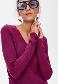 Sweter w serek w ciemnopurpurowym prążkowanym dzianinie, z długimi rękawami. Uzupełniony dużymi okularami w kolorze żółwim i okrągłymi kolczykami.