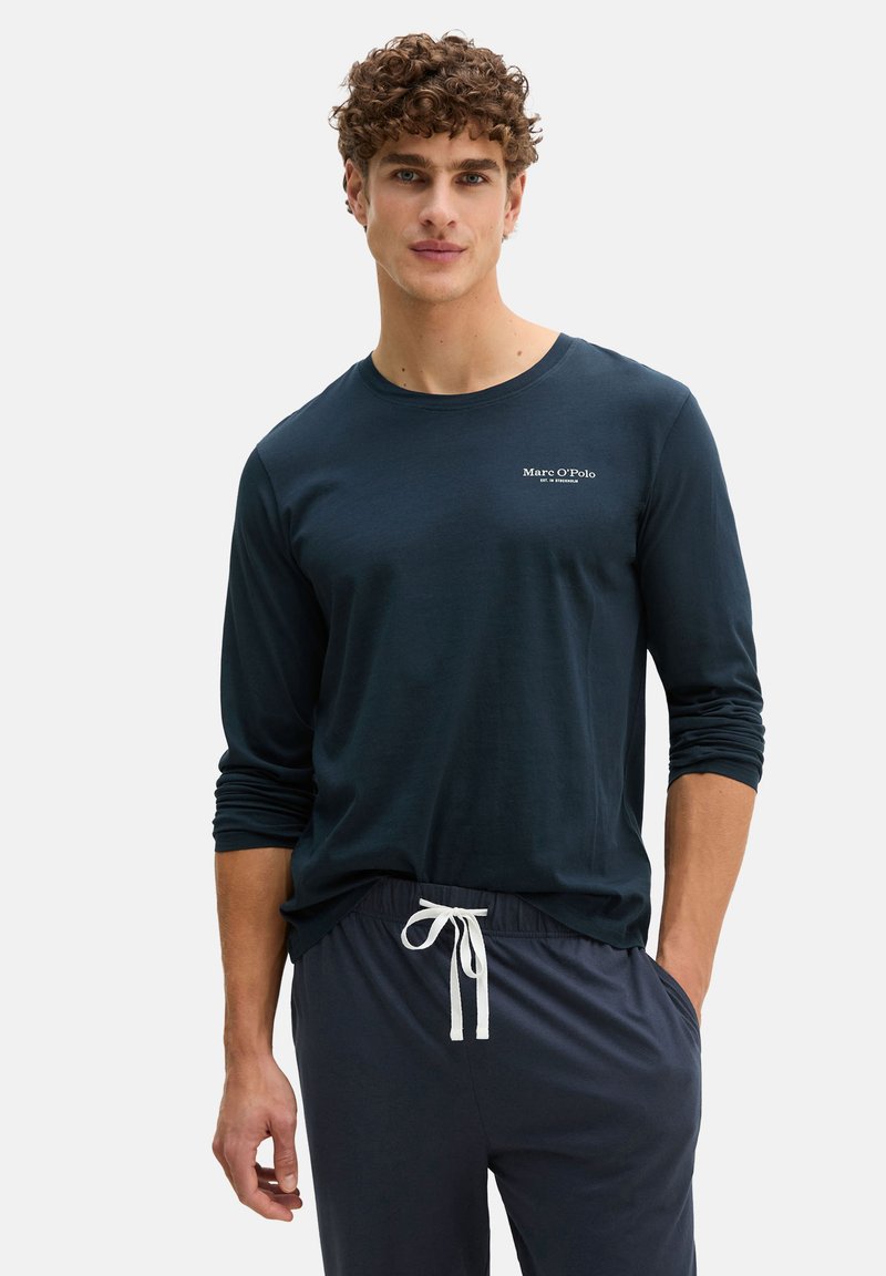 Junger Mann mit lockigem Haar, der ein marineblaues Langarmshirt und passende Hosen mit Kordelzug trägt und mit einer Hand in der Tasche steht.