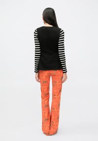 Juodas be rankovių megztinis ant dryžuotos ilgomis rankovėmis palaidinės, derinamas su oranžinėmis raštuotomis kelnėmis su rudos spalvos akcentais.