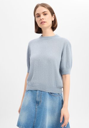 Jeune femme debout portant un pull en maille bleu clair à manches courtes et une jupe en jean bleu délavé sur fond blanc.