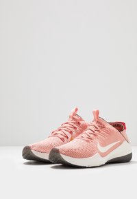 Rosa Nike träningsskor med en strukturerad stickad överdel och vit swoosh, placerade mot en enkel bakgrund. Snören och dragflikar synliga.