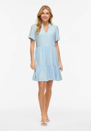 Jeune femme debout, portant une robe courte bleu clair à manches courtes jusqu'aux genoux en tissu ajouré et des sandales blanches, souriant à la caméra.