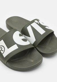 Sandales glissantes vert olive avec une large bride portant des lettres blanches. Semelle texturée pour l'adhérence et le confort. Matériau synthétique lisse.
