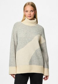 Heledhalli ja kreemjas kampsun kõrge kaelusega ja soonikuid imiteeriva tekstuuriga, millel on ees suur kreemjas abstraktne muster ja pikad varrukad.