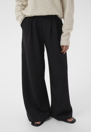 Person wearing wide-leg black trousers, beige ribbed long-sleeve sweater, and black open-toe sandals standing against a plain background.