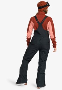 Pantalones de nieve negros con tirantes ajustables, combinados con una parte superior de forro polar en tonos burdeos y rosa. Aberturas con cremallera en las piernas y gorro de punto texturizado.