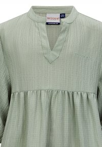 Hellgrüne Tunika aus strukturiertem Stoff, mit V-Ausschnitt und geraffter Taille. Mit "Retour Jeans"-Stickerei auf der Brust.