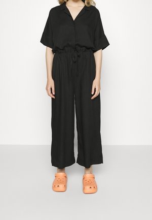 Czarny kombinezon lniany z kołnierzykiem, krótkimi rękawami i sznurkiem w talii. Do tego jasnopomarańczowe chodaki z otworami wentylacyjnymi.