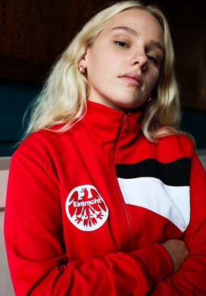 Rode zip-up jas met zwart-witte accenten, met een cirkelvormig Eintracht-logo. Soepele stof met een sportief ontwerp en hoge kraag.