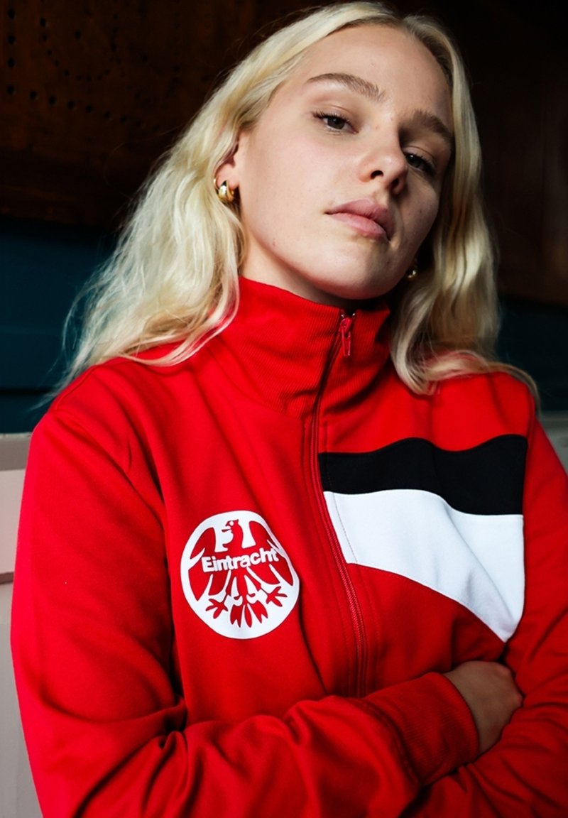 Rode zip-up jas met zwart-witte accenten, met een cirkelvormig Eintracht-logo. Soepele stof met een sportief ontwerp en hoge kraag.