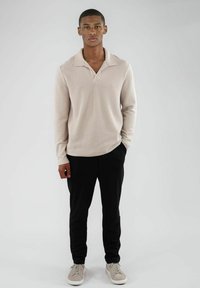 Beige ribbestrikket langermet polo med krage, kombinert med svarte bukser og lysegrå sneakers, står på en ensfarget bakgrunn.