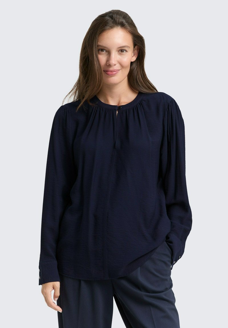 Blouse marine à manches longues avec un col rond, des détails froncés et une fermeture à bouton au cou. Tissu lisse avec une légère texture.