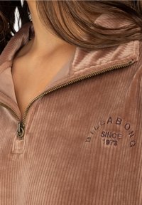 Hnědý kordový pulovr na polo zip s ribbovanou texturou. Obsahuje zip a vyšívaný detail "Billabong Since 1973" v tmavě fialové barvě.