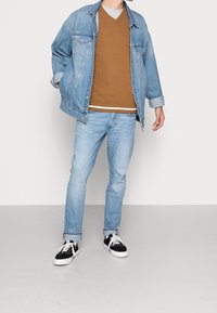 Ljusblå denimjacka över en brun tröja med en grå undertröja, kombinerat med ljusblå jeans och svarta sneakers med vita sålar.