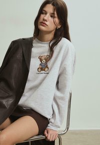 Grå sweatshirt med en broderet teddybjørn iført en stribet skjorte og teksten "Jeg kan ikke bære mandag", parret med en brun miniskirt.