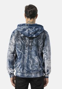 Blauwe en witte hoodie met een gestructureerd, vervaagd patroon. Heeft grafische details en een tekening van vleugels op de achterkant.